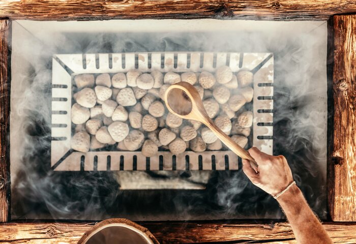 Eine Hand hält einen Holzlöffel über einen Grill mit glühenden Kohlen, umgeben von Rauch. Der Grill ist aus Holz und hat eine rechteckige Form. Die Atmosphäre wirkt rustikal und einladend.