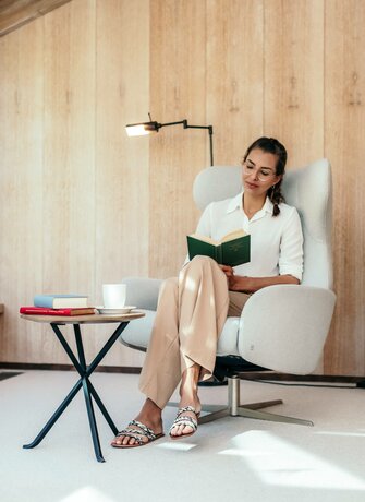 Eine Frau liest in einem Sessel neben einem Beistelltisch mit Büchern und Tasse in der Bibliothek.