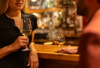 Eine Frau hält ein Weinglas an der Bar, während ein Gast mit einem Glas daneben steht.