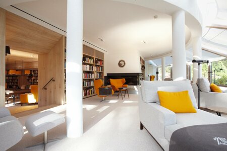 Heller Aufenthaltsbereich mit weißen Sesseln, orangefarbenen Stühlen, Bücherregalen und Blick in einen offenen Bibliotheksraum.