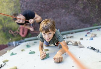 Ein Kind klettert an einer Kletterwand, während eine zweite Person es mit Seil sichert.