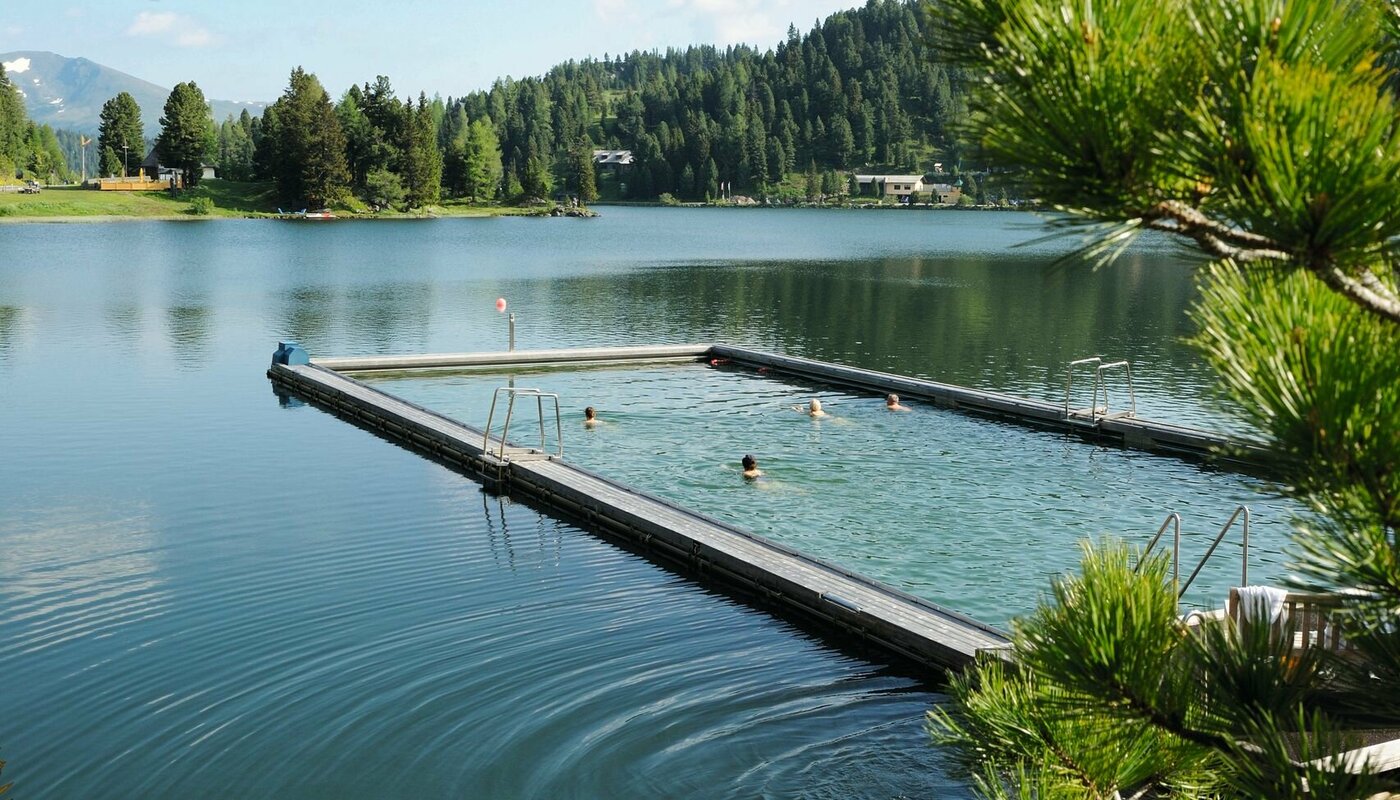 Ein beheiztes See-Bad liegt im Turracher See, mit Steg, ruhigem Wasser und bewaldeten Bergen im Hintergrund.
