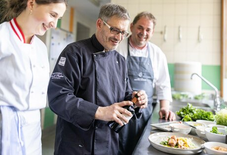 Ein Koch und zwei Teammitglieder richten in der Hotelküche mehrere Teller mit Speisen an.