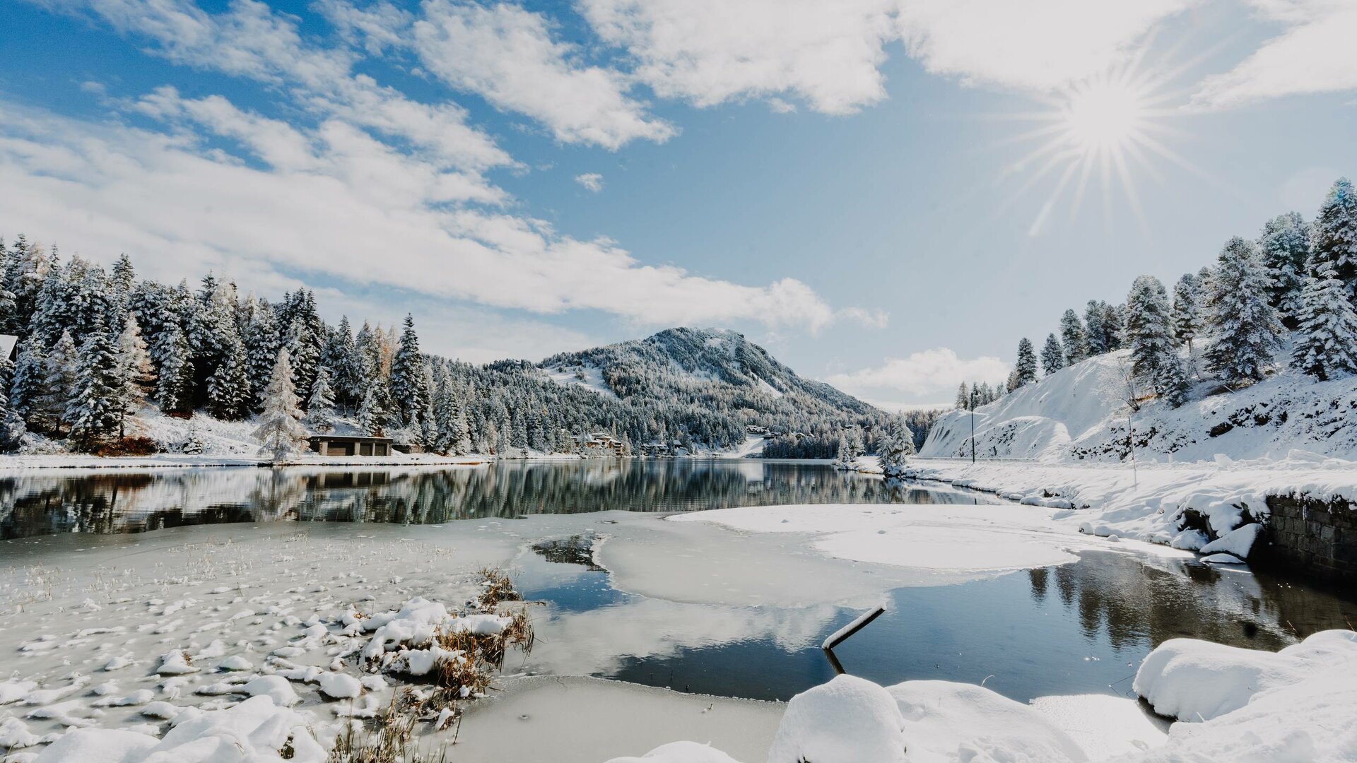 Der verschneite Turracher See liegt ruhig unter sonnigem Winterhimmel, mit Wäldern und Bergen am Ufer.