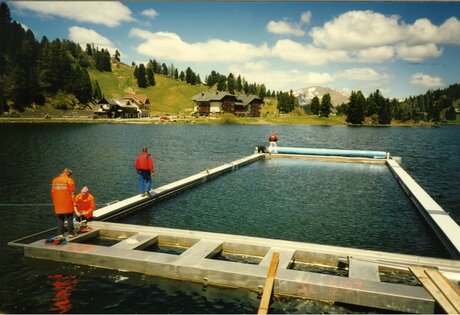 Guests stand on the dock at the heated lake pool in Lake Turracher See, with alpine meadows and forest behind.