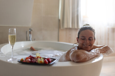 Eine Frau entspannt in der Badewanne eines Zimmers, daneben stehen ein Tablett mit Frühstück und ein Glas Sekt.