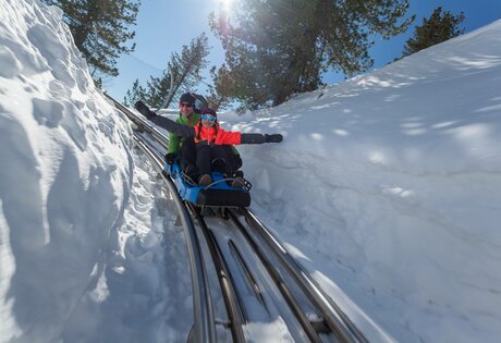 Zwei Personen fahren mit einer Rodel durch eine schmale Schneerinne unter blauem Himmel.