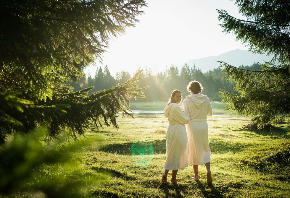 Zwei Personen stehen im Morgenlicht auf einer Wiese am See, umgeben von Bäumen und mit Blick in die Berge.