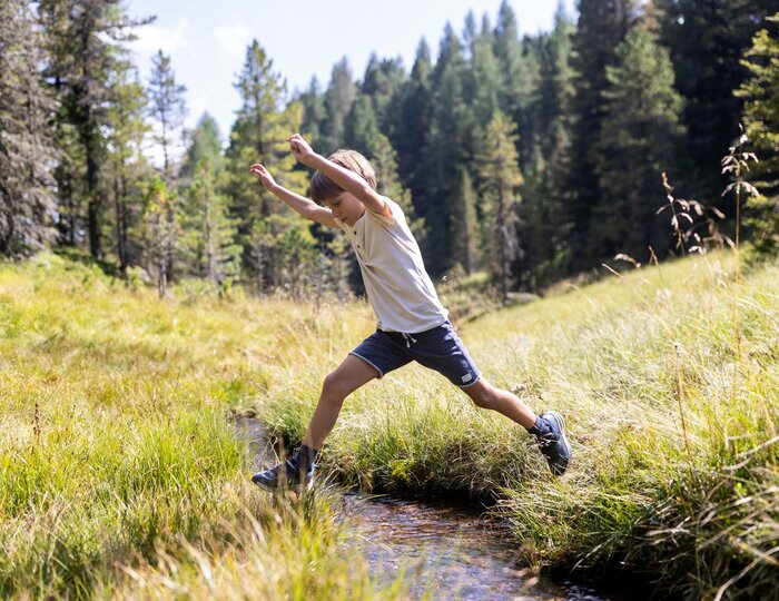 Ein Kind springt mit ausgestreckten Armen über einen kleinen Bach auf einer Wiese im Wald.