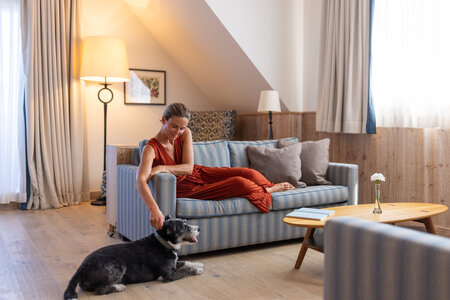 Ein Hund liegt vor dem Sofa in einem Hotelzimmer mit Lampe, Vorhängen und Holzwandverkleidung.