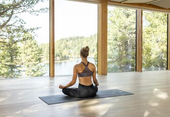 Eine Frau sitzt auf der Yogamatte vor den großen Fenstern mit Blick auf den See und den Wald.