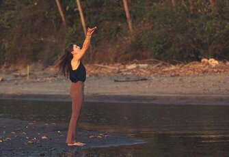 A woman stands at the water’s edge with her arms raised in a yoga pose.