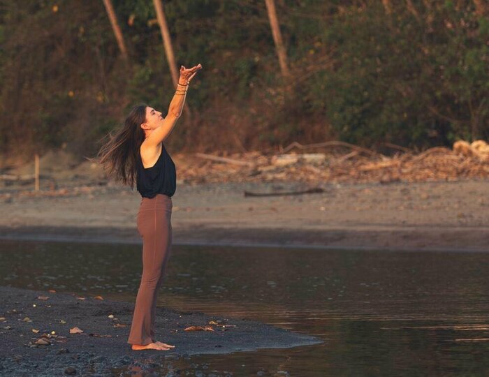 Eine Frau steht am Seeufer und hebt die Arme in einer Yoga-Pose.