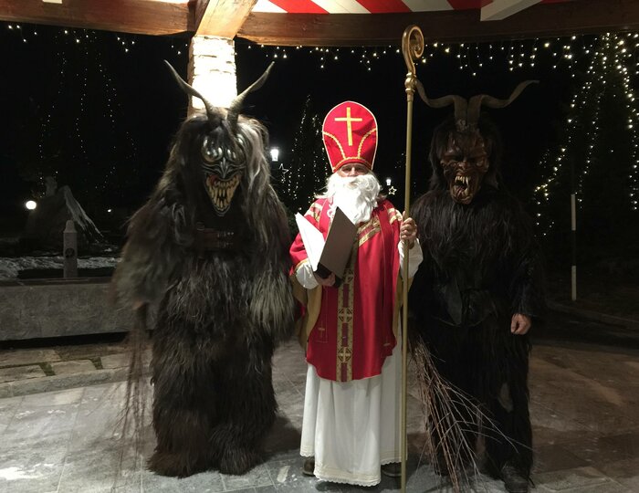 Nikolaus steht zwischen zwei Krampussen unter Lichterketten im nächtlichen Freien.