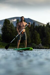 Ein Mann steht barfuß auf einem SUP-Board auf dem See, im Hintergrund liegen Wald und Berge.