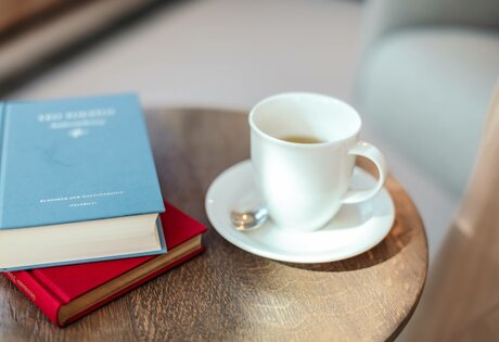 Auf einem Holztisch stehen ein weißer Kaffeebecher auf Untertasse und mehrere Bücher, darunter ein blaues und ein rotes.