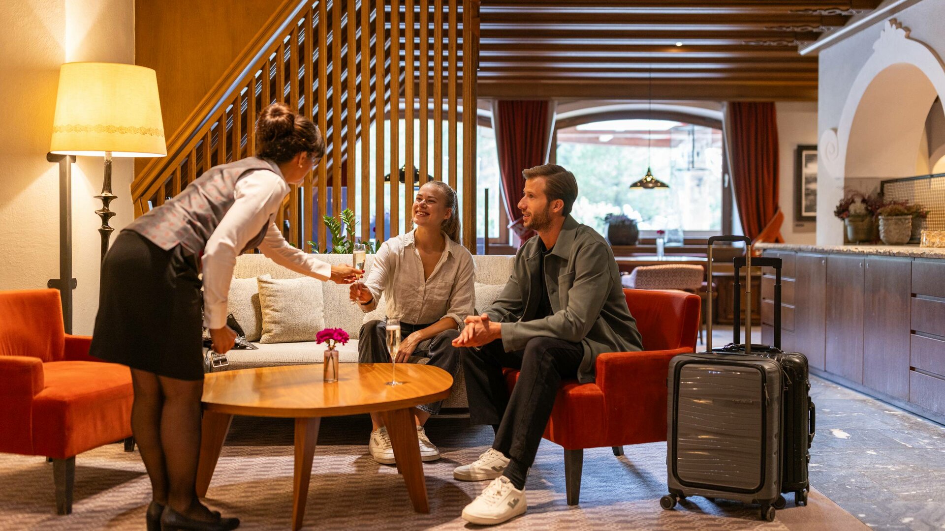 Eine Mitarbeiterin begrüßt zwei Gäste in der Lounge des Hotels, neben ihnen stehen Koffer.
