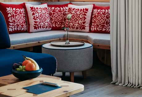 Ein gemütlicher Sitzbereich im Hotelzimmer mit blauer Sesselfläche, Rundtisch und Sitzbank mit roten Kissen am Fenster.