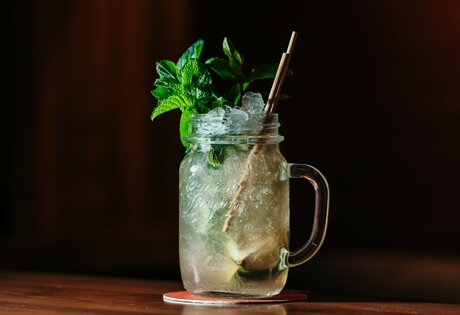 Ein Cocktail im Glas mit Minze, Eis und Strohhalm steht auf einem Holztisch.