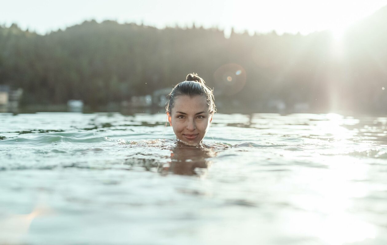 Eine Frau schwimmt im beheizten See-Bad, im Hintergrund liegen Wald und Berge im Licht der tiefen Sonne.