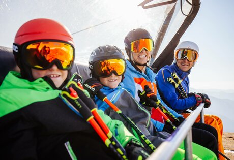 Four skiers sit on a chairlift, wearing helmets and ski goggles.