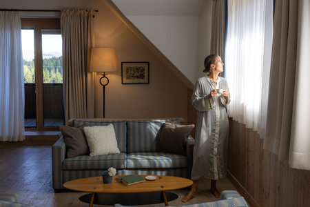 Eine Frau steht im Wohnbereich einer Suite am Fenster und blickt hinaus; Sofa, Stehlampe und Couchtisch sind zu sehen.
