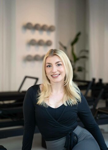 Pilates im Hotel Hochschober mit Trainerin Eine lächelnde Trainerin sitzt in einem Pilatesraum mit Geräten und zeigt eine entspannte, aufrechte Haltung.