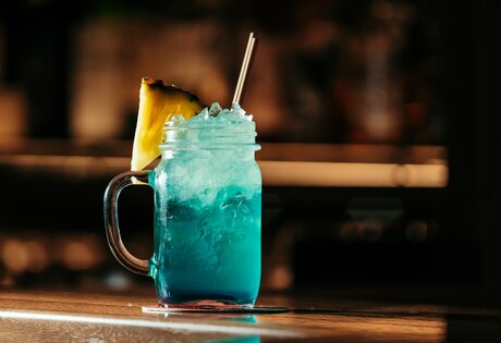 Ein türkisblauer Cocktail im Glas mit Ananasscheibe und Strohhalmen steht auf der Bar.
