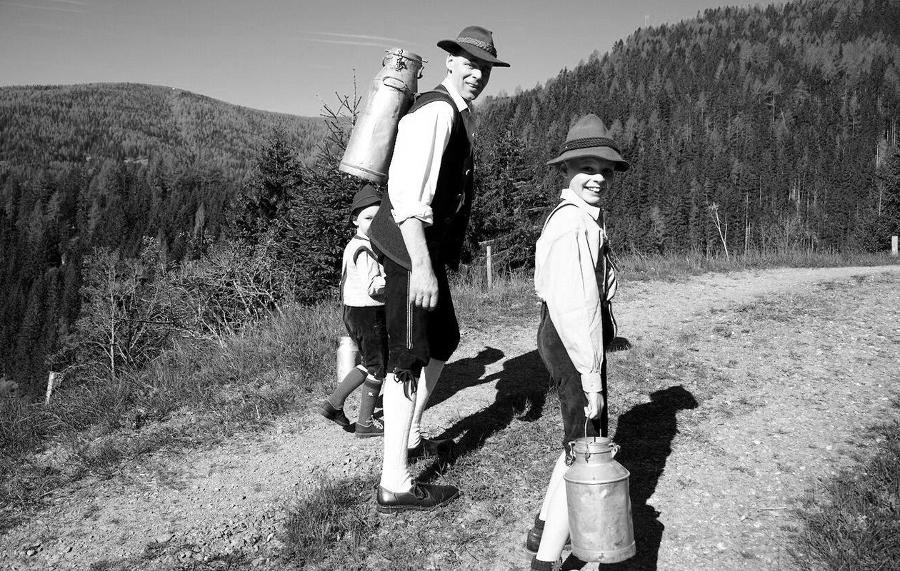 Zwei Wanderer mit Hüten tragen Milchkannen auf einem Bergpfad in der Landschaft.