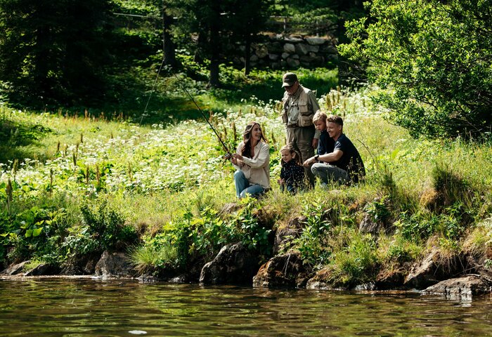 Eine Familie sitzt und steht am grünen Ufer am Wasser und genießt einen Sommerausflug in der Natur.