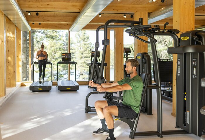 Ein Mann trainiert an einer Kraftmaschine, während eine Frau auf dem Laufband im hellen Fitnessraum trainiert.