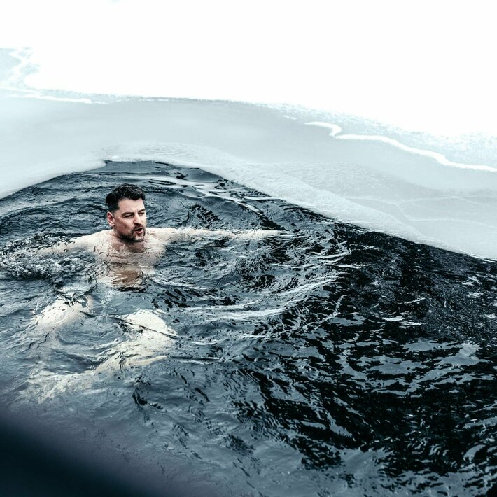 Ein Mann schwimmt in einem dunklen, eisigen See-Bad unter Schnee am Ufer.