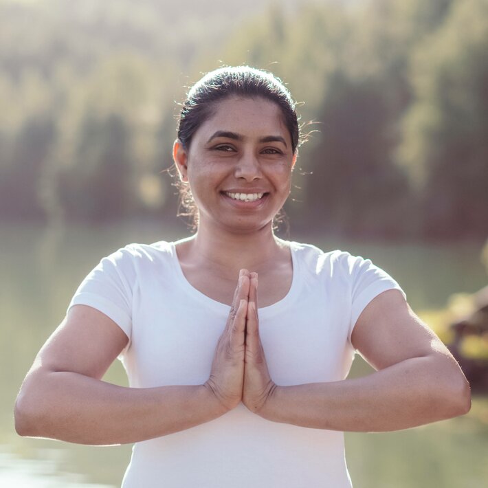 Eine Frau steht im Yoga-Gruß am Seeufer und lächelt in die Kamera.