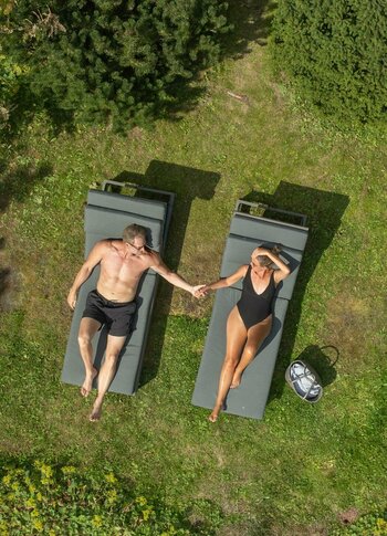 Zwei Personen liegen auf Gartenliegen im Gras und halten sich an den Händen, umgeben von Wiesen und Büschen.
