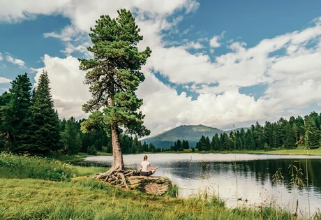 A person sits on the lakeshore in front of mountain scenery and wooded slopes.
