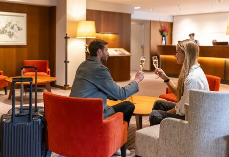 Ein Paar stößt in der warm gestalteten Hotel-Lounge mit Gepäckwagen an.