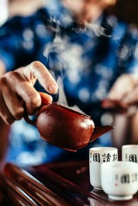 Eine Hand gießt Tee aus einer kleinen Kanne in eine Tasse; im Vordergrund stehen weitere Teebecher mit Schriftzeichen.