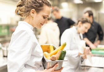 Eine Auszubildende trägt frisches Gemüse in der Hotelküche, während im Hintergrund weitere Mitarbeitende arbeiten.
