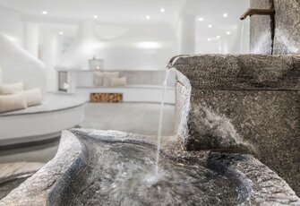 Eine Steinschale mit fließendem Wasser steht in einem hellen Wellnessbereich mit Liegen und geschwungener Sitzbank.