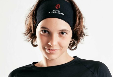 Eine Frau trägt ein schwarzes Hotel-Hochschober-Shirt und ein passendes Stirnband vor hellem Hintergrund.