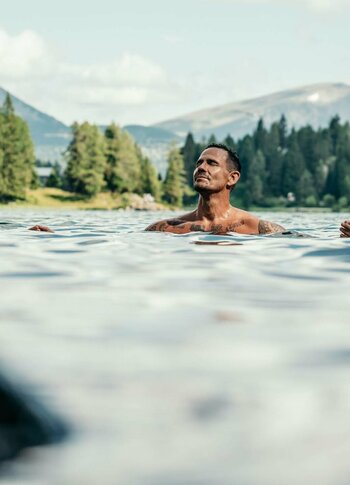 Ein Mann schwimmt im kalten Bergsee, umgeben von Nadelwald und Bergen unter wolkigem Himmel.