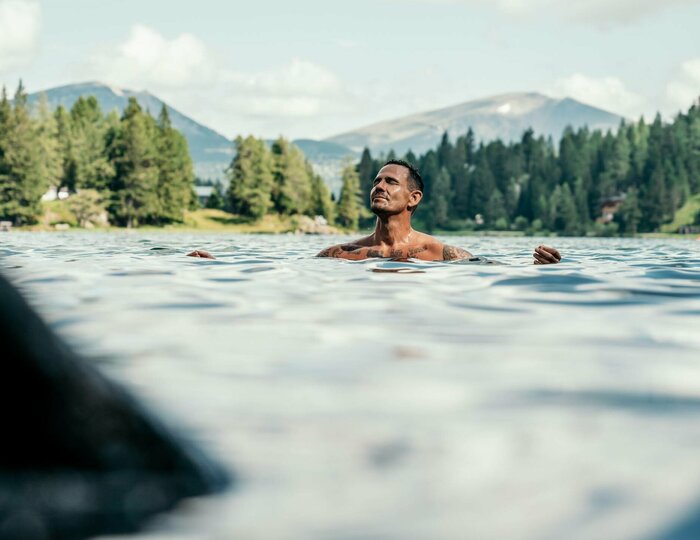 Ein Mann schwimmt im kalten Bergsee, umgeben von Nadelwald und Bergen unter wolkigem Himmel.