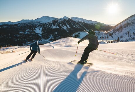 Two skiers descend a freshly groomed slope with snowy mountains and low sun in the background.