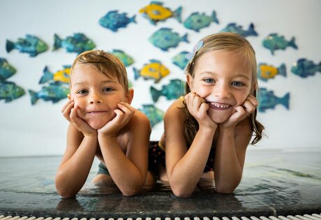 Two children are lying in the indoor pool and smiling at the camera, with colorful fish shapes on the wall behind them.