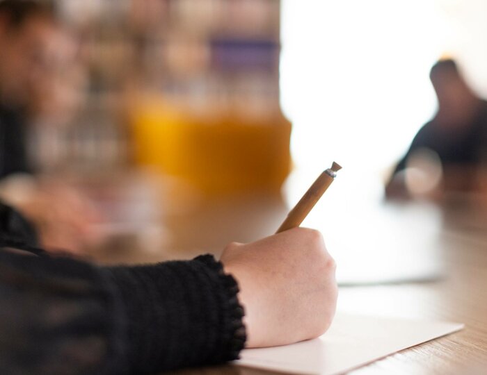 A person takes notes during a job interview at a table, with two blurred people in the background.