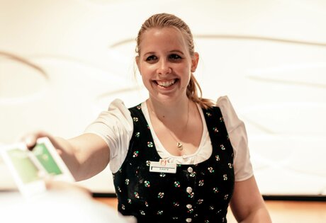 Eine Mitarbeiterin lächelt und reicht im Hotelservice eine Karte nach vorn.