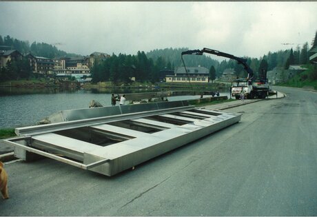 A large structural element lies on the shore of Turracher See, with a crane truck parked beside the road.