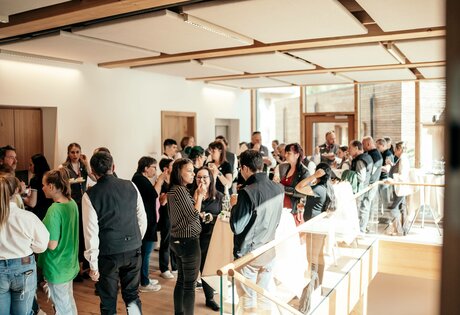 Gäste stehen bei einem Seminar-Empfang in einem hellen Raum mit Holzdecke und Glasgeländer zusammen.