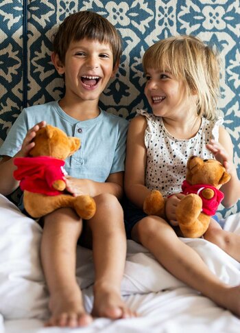 Kinder auf dem Hotelbett im Familienzimmer Zwei Kinder sitzen lachend auf dem Bett in einem hellen Hotelzimmer und halten Plüschtiere in den Händen.
