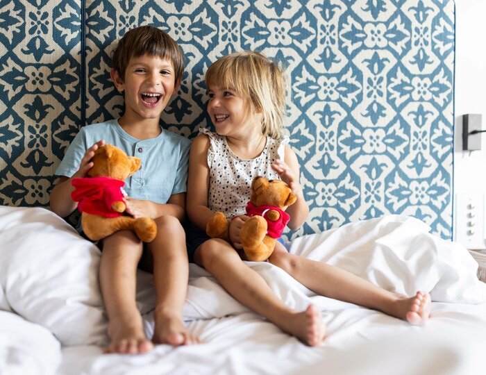Kinder auf dem Hotelbett im Familienzimmer Zwei Kinder sitzen lachend auf dem Bett in einem hellen Hotelzimmer und halten Plüschtiere in den Händen.